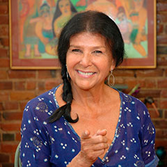 Photographie couleur d'une femme souriante, portant une chemise bleue à motifs blancs. Elle pointe le doigt vers la caméra. Ses cheveux noirs sont tressés.
