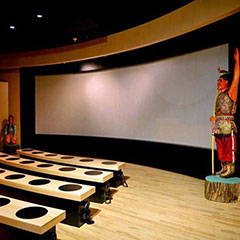 Colour photograph of a screening room: a giant screen, some benches and two sculptures representing two Abenakis.