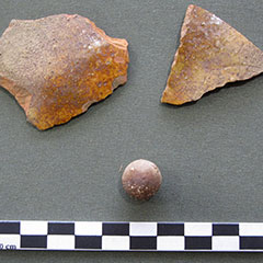 Photographie couleur de deux tessons de terre-cuite de couleur orangée et une balle de fusil en plomb.