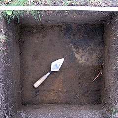 Colour photograph of an archeological test pit (squared-shape hole) where a blackened area and a trowel are shown.