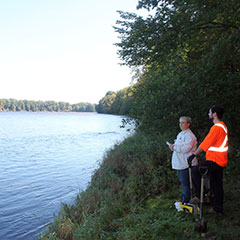 Colour photograph of a man and a woman near a river, each holding a shovel.