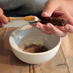 Close-up colour photograph of hands cleaning metal artifact with a toothbrush over a porcelain bowl.