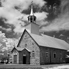 Photographie noir et blanc d'une église en pierre et de son clocher. On voit également de gros nuages blancs.