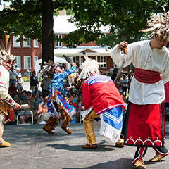 Photographie couleur de quatre danseurs portant des vêtements traditionnels de cuir ou de tissu. Tous portent également des mocassins et une coiffe en plumes sur la tête.