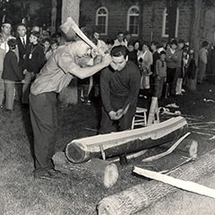 Black and white photograph of two men beating a tree trunk with the back of their axes. A crowd watches them.